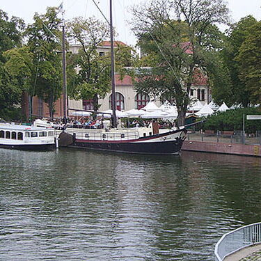 Zwei Boote liegen am Ufer eines Flusses in Köpenick, umgeben von grünen Bäumen und einem Gebäude mit weißen Sonnenschirmen.