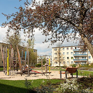 Ein Spielplatz mit bunten Klettergeräten liegt in einem grünen Innenhof, umgeben von mehrstöckigen Wohngebäuden.