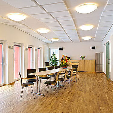 Ein heller Festsaal mit Holzboden und einem langen Tisch, umgeben von Stühlen, dekoriert mit einem Blumenarrangement.