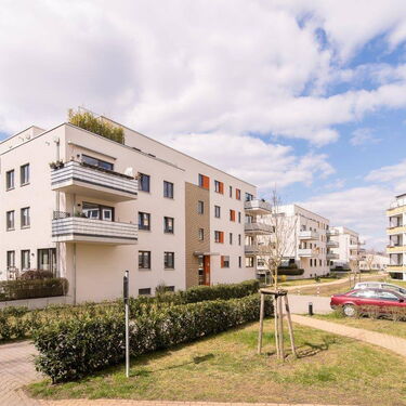 Mehrfamilienhäuser mit Balkonen und geparkten Autos in einer sonnigen Wohnanlage unter blauem Himmel.