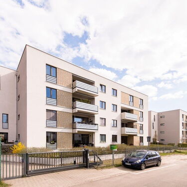 Moderne Wohngebäude mit Balkonen und einem Parkplatz, umgeben von einer gepflegten Landschaft und einer ruhigen Straße.