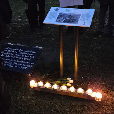 Kerzen und Blumen schmücken eine Gedenktafel bei einer Abendveranstaltung auf einem historischen Gelände.