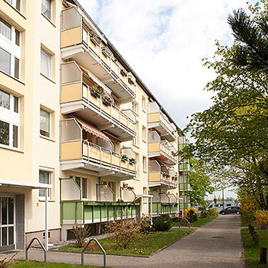 Mehrstöckige Wohngebäude mit Balkonen und gepflegten Grünanlagen säumen einen Fußweg unter blauem Himmel.