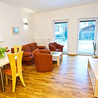 Helles Wohnzimmer mit Holzboden, gemütlicher Sitzecke, Esstisch und Blick auf den Garten durch große Fenster.