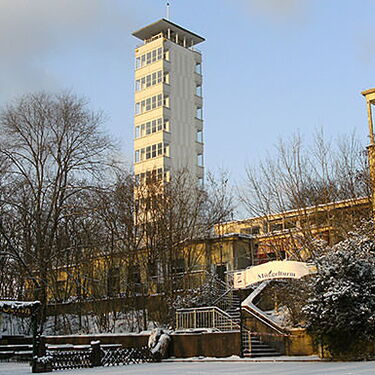 Ein verschneiter Aussichtsturm in Köpenick erhebt sich vor winterlichen Bäumen und einem historischen Gebäude.