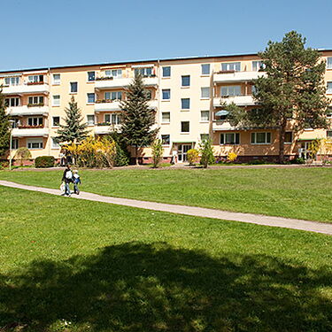 Ein mehrstöckiges Wohngebäude mit Balkonen steht vor einem großen grünen Rasen, auf dem zwei Menschen spazieren.