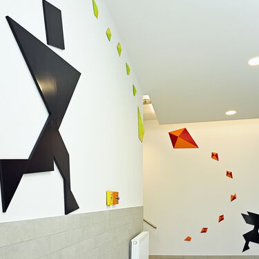 Eine stilisierte schwarze Figur läuft an einer Wand, umgeben von bunten geometrischen Formen in einem modernen Raum.