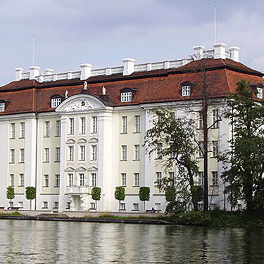 Ein historisches Schloss mit rotem Dach spiegelt sich malerisch im ruhigen Wasser von Köpenick wider.