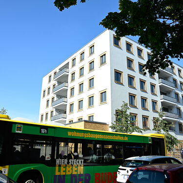 Ein Stadtbus fährt vorbei an einem modernen Wohngebäude mit Balkonen und Bäumen in der Umgebung.