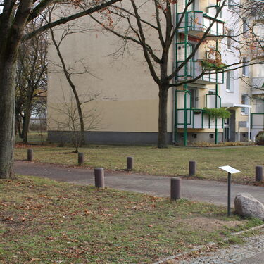 Ein Wohngebäude mit Balkonen und herbstlichen Bäumen steht neben einem gepflasterten Weg in einer ruhigen Nachbarschaft.