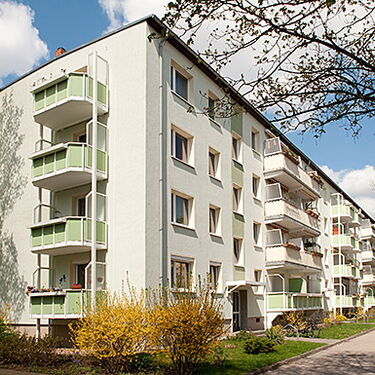 Ein mehrstöckiges Wohngebäude mit grünen Balkonen steht bei sonnigem Wetter in einer baumreichen Straße.