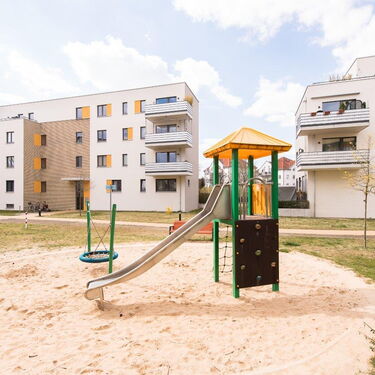 Ein moderner Spielplatz mit Rutsche und Klettergerüst liegt zwischen drei weißen Wohngebäuden unter blauem Himmel.