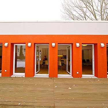 Eine moderne Festsaal-Fassade mit leuchtend orangefarbenen Akzenten und großen Fenstern öffnet sich zu einem Holzbodenbereich.