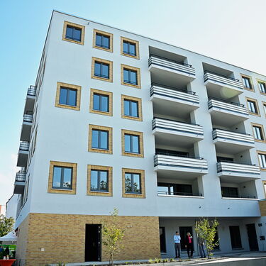 Ein modernes Wohngebäude mit mehreren Balkonen und großen Fenstern steht unter einem klaren Himmel, während Besucher davor stehen.